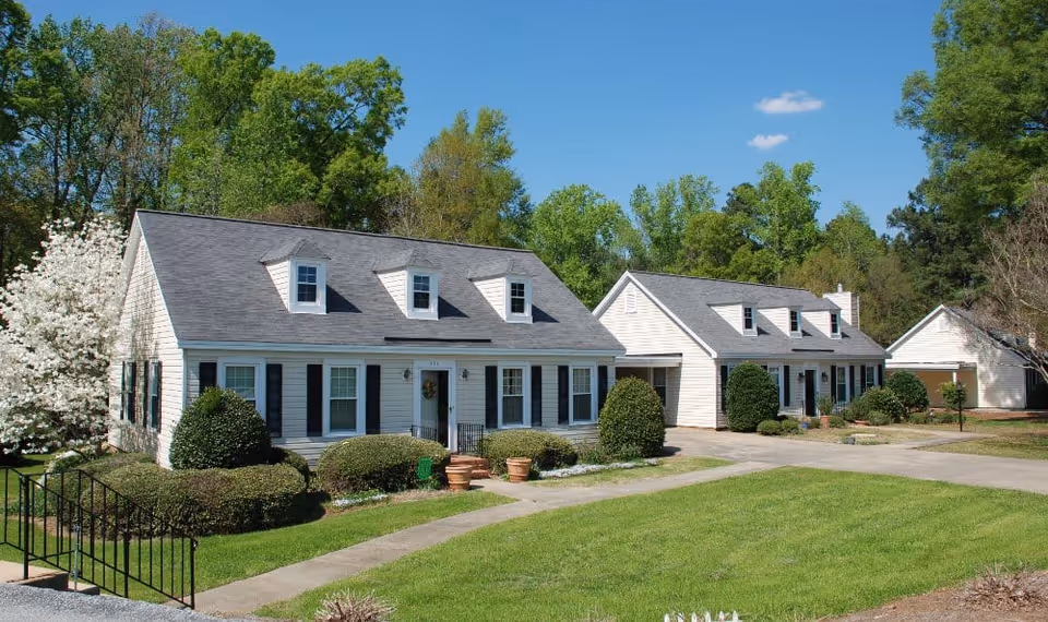 Two white residential-style buildings with black shutters and gray roofs surrounded by green trees and bushes under a clear blue sky.