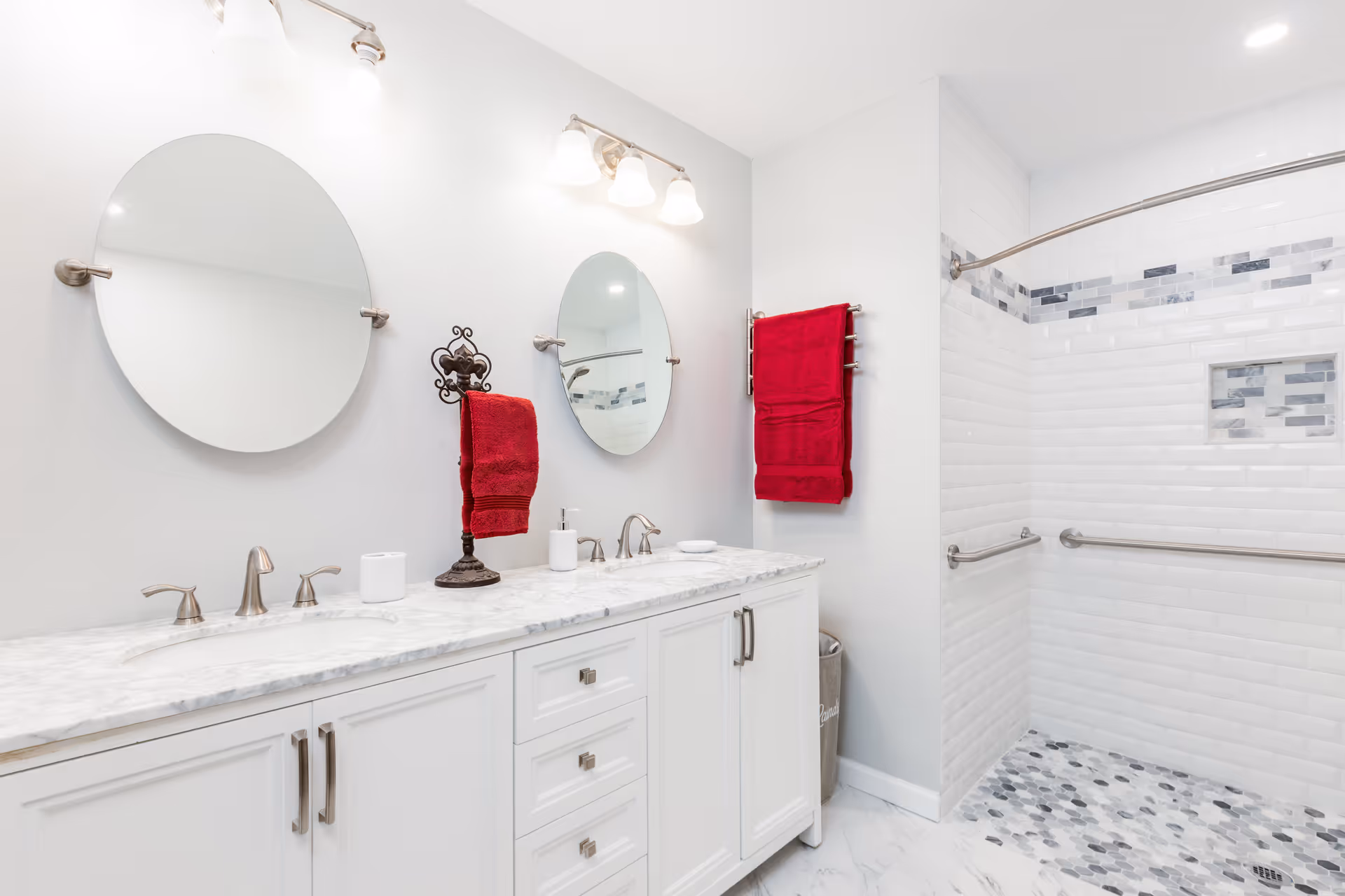 Bright and clean bathroom with white cabinetry and marble countertop featuring two sinks and two round mirrors above. The shower area has white subway tiles with a decorative strip of gray and blue tiles, a built-in niche, and stainless steel grab bars. Red towels hang on a towel rack and a decorative stand.