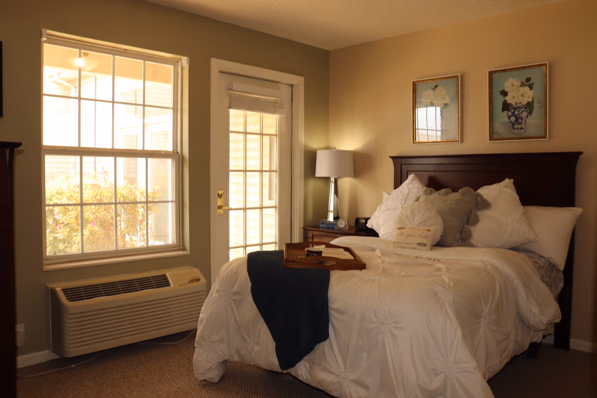 A cozy bedroom with a neatly made bed featuring white bedding and multiple pillows. There is a wooden headboard and a bedside table with a lamp, books, and a clock. Two framed floral pictures hang on the wall above the bed. A window and a glass door allow natural light into the room, and an air conditioning unit is installed below the window.