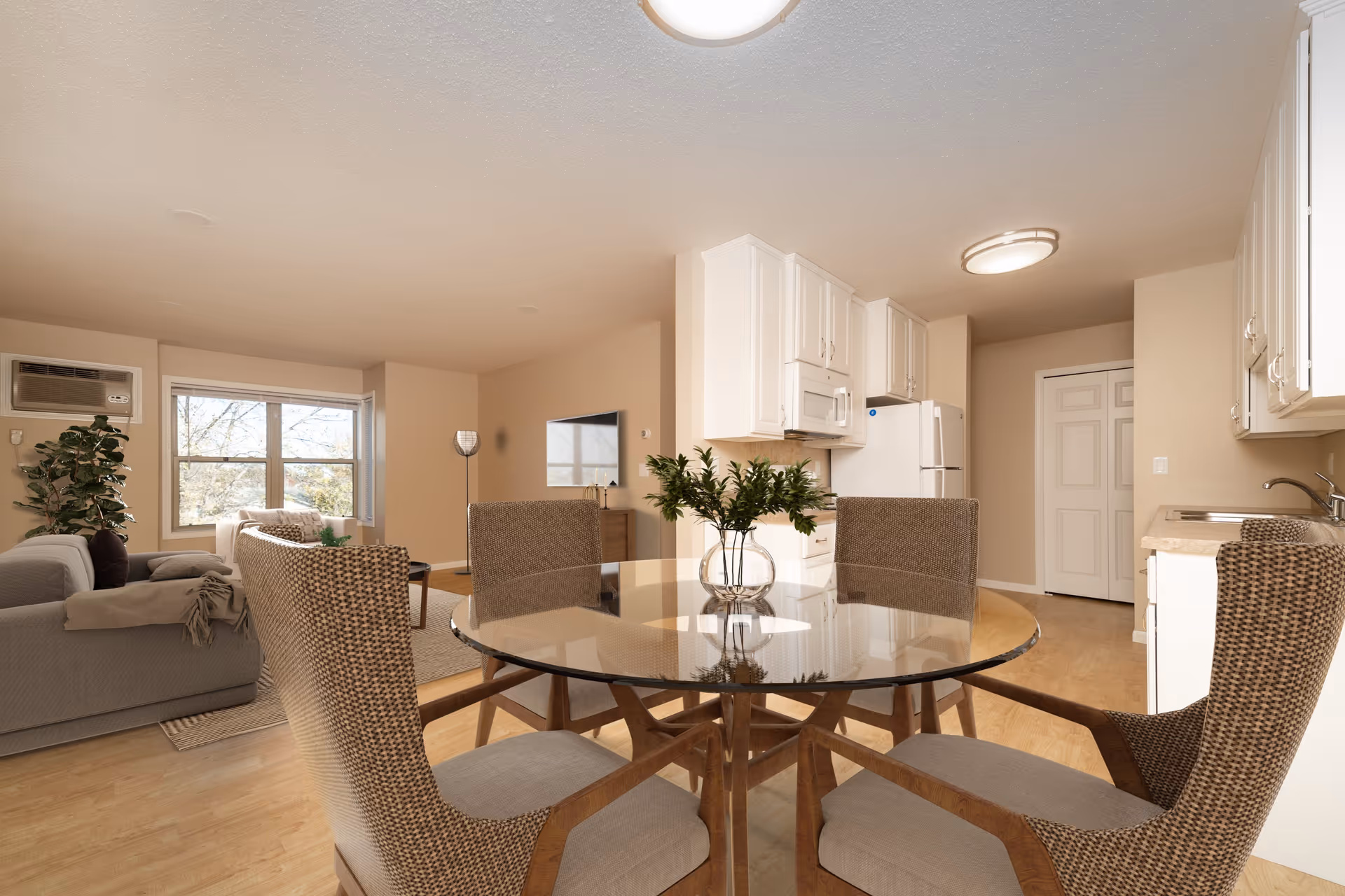 Interior view of a senior living facility showing a combined dining and living area. In the foreground, there is a round glass dining table with four cushioned chairs. The background features a living room with a gray sofa, a window with an air conditioning unit, a TV mounted on the wall, and a kitchen area with white cabinets, a refrigerator, microwave, and sink. The walls are beige and the floor is light wood.