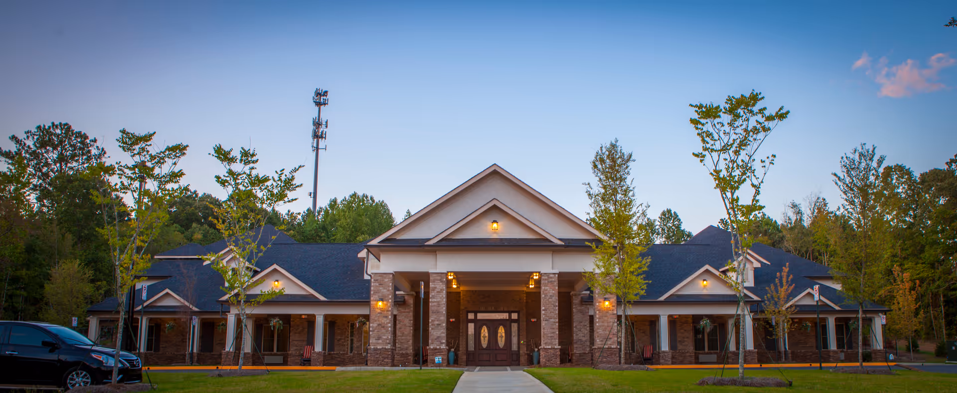 Front exterior view of a single-story senior living facility building with a peaked roof, brick and stone facade, illuminated porch lights, young trees planted in front, and a car parked on the left side.