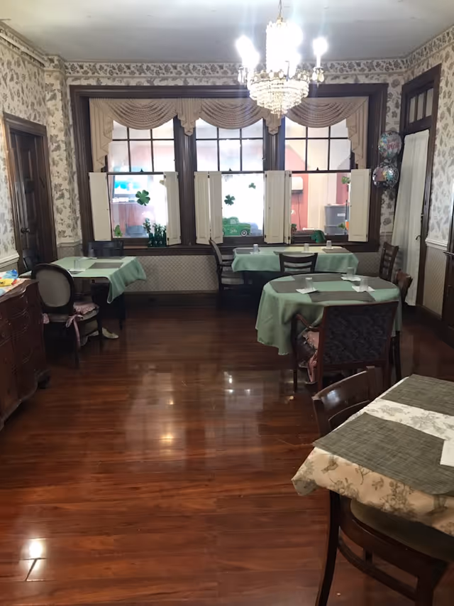 A dining room with wooden floors and floral wallpaper. Several tables are covered with green and beige tablecloths, each set with placemats and napkins. There are wooden chairs around the tables. Large windows with decorative valances and shutters let in natural light. A chandelier hangs from the ceiling, and there are some St. Patrick's Day decorations on the windows.