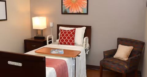 A cozy bedroom in a senior living facility featuring a single bed with white and red bedding, a bedside table with a lamp, a tray table with magazines, a patterned armchair with a beige pillow, and a framed flower artwork on the wall.