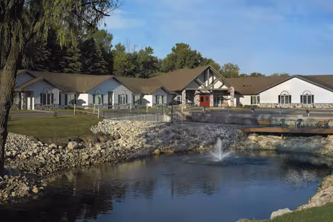 Single-story senior living building with a pond and fountain in front, lawns and trees surrounding it.
