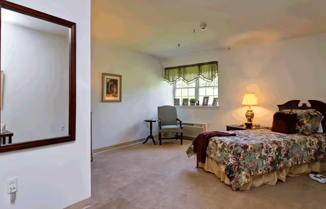 A bedroom in a senior living facility with a bed covered in floral bedding, a nightstand with a lamp, a chair near a window with green valance curtains, and a large mirror on the wall.