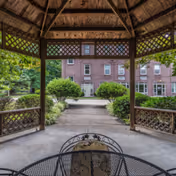 View from inside a wooden gazebo looking out towards a brick building with multiple windows. The gazebo has lattice panels and a metal table with chairs in the foreground. Surrounding the gazebo are green bushes and trees.