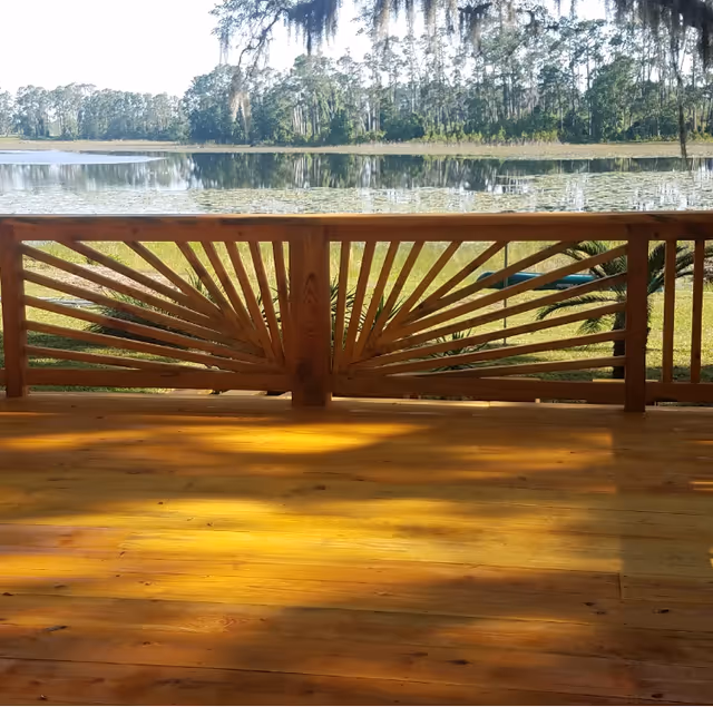 View from a wooden deck with a decorative railing overlooking a calm lake surrounded by trees and greenery under a clear sky.