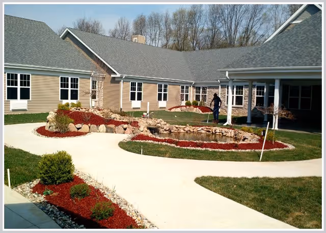 Outdoor courtyard area of a senior living facility with a landscaped garden featuring a small pond surrounded by rocks and red mulch. A person is tending to the garden near the pond. The building has beige siding, multiple windows, and a gray shingled roof. Trees are visible in the background.