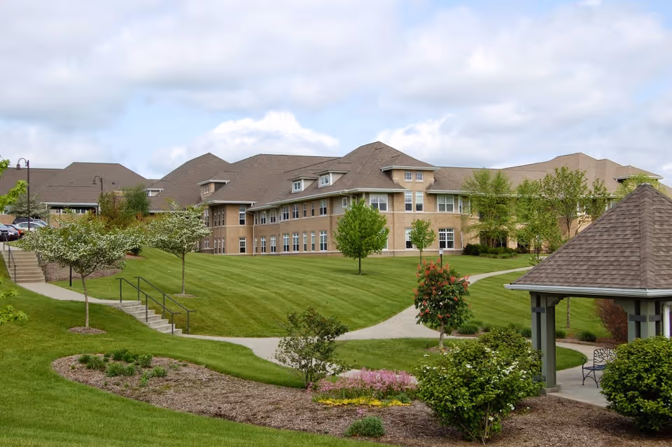 Well-maintained brick senior living building with manicured lawns, walking paths, trees, and a gazebo.
