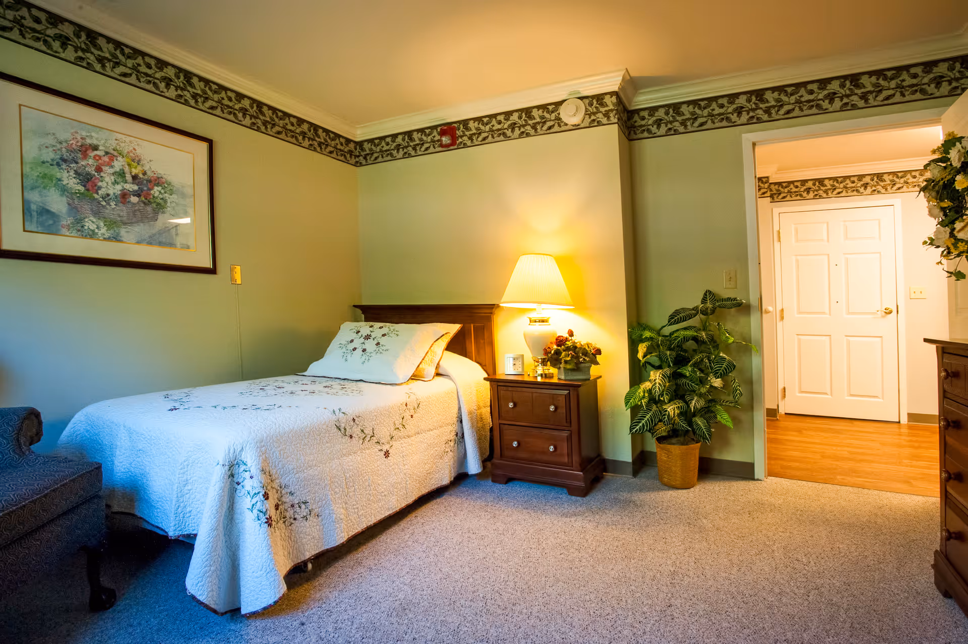 A cozy bedroom in a senior living facility featuring a single bed with a floral quilt and matching pillow, a wooden nightstand with a lamp and a small plant, a framed floral painting on the wall, a potted plant near the doorway, and a glimpse of a hallway with white doors and wooden flooring.