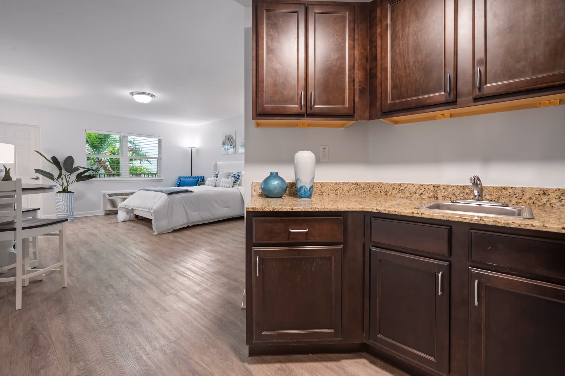Open studio interior showing a kitchenette with dark cabinets and granite countertop opening to a bed, small dining area, and window.