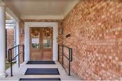 Entrance to a building with double wooden doors featuring decorative glass panels. The doors are framed by a brick wall and have two black handrails on either side of the steps leading up to the entrance. There are three black mats on the concrete floor in front of the doors.