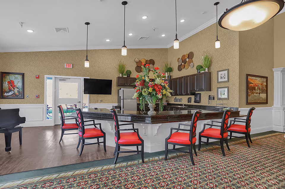 Bright senior living common area with a large island bar, red-upholstered chairs, pendant lights, and a floral centerpiece.