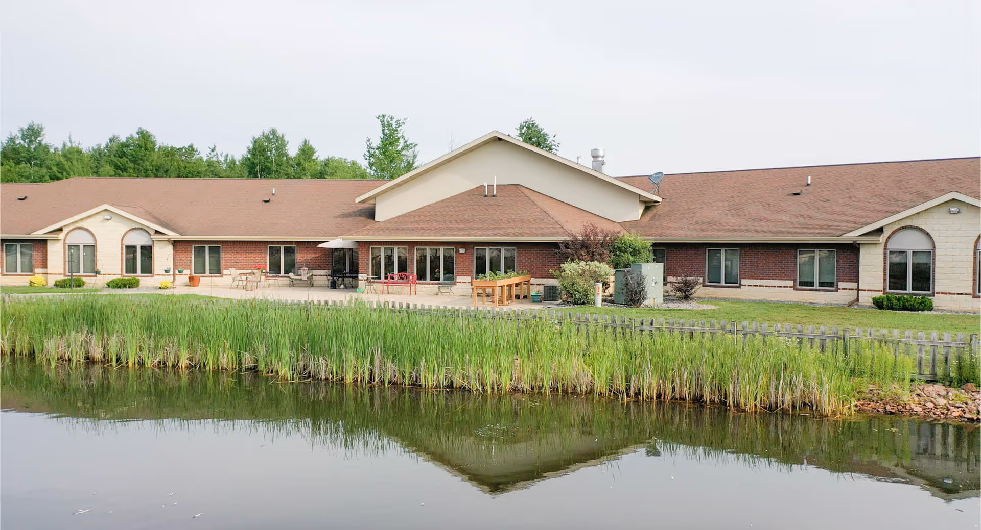 Single-story senior living building with a patio and outdoor seating facing a pond with tall reeds and its reflection.