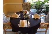 A round dining table set for four with a black tablecloth, white folded napkins, silverware, glasses, a small floral centerpiece, and a menu, surrounded by upholstered chairs with floral patterns, next to a window with green plants visible outside.