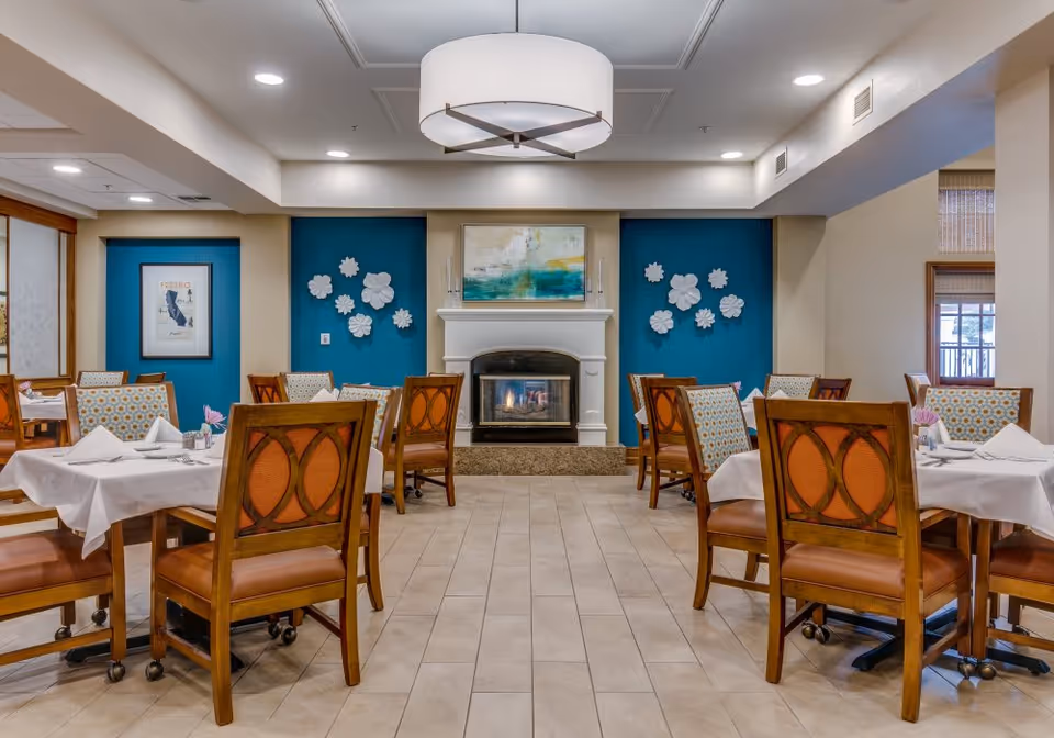 A dining room with several wooden tables and chairs arranged neatly. Each table is covered with a white tablecloth and set with napkins and utensils. The room features a fireplace in the center of the far wall, flanked by blue accent walls decorated with white floral wall art. Above the fireplace is a framed abstract painting. The ceiling has recessed lighting and a large circular light fixture.