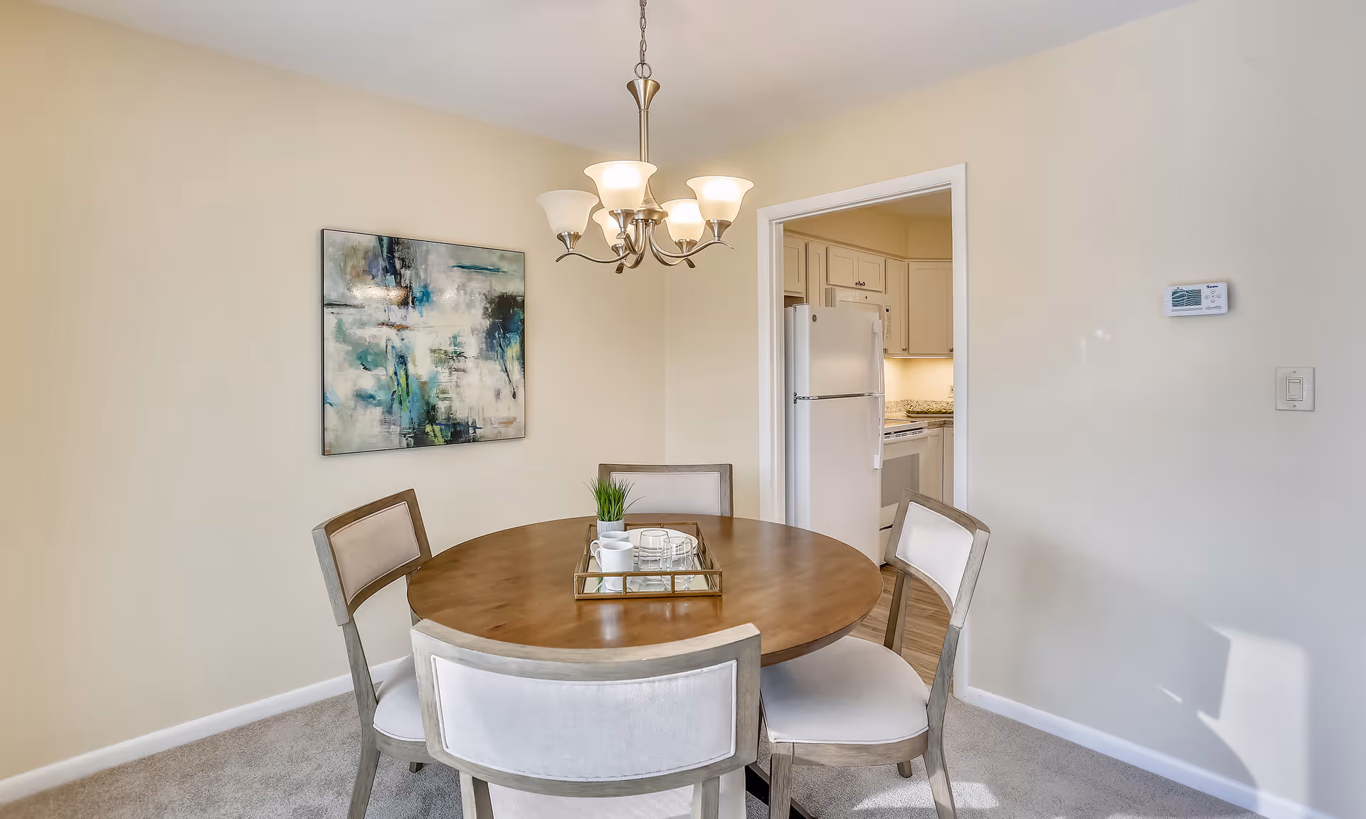 Round wooden dining table with four upholstered chairs under a chandelier, a wall painting, and a view into an adjacent kitchen.