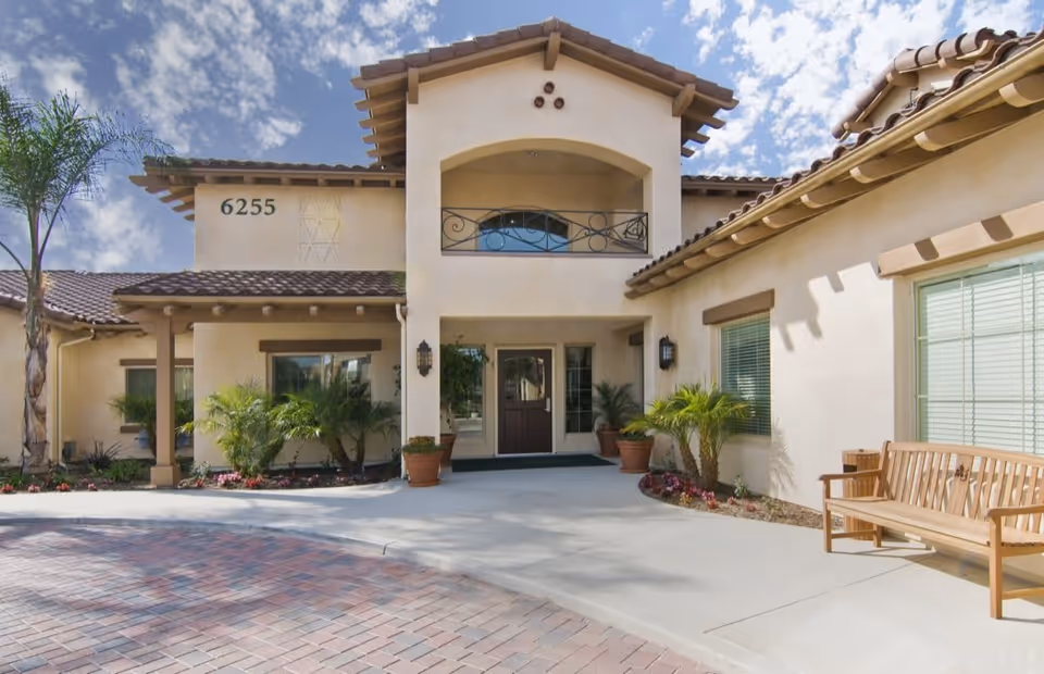 Exterior front entrance of a building with beige stucco walls and a tiled roof, featuring a covered porch area with potted plants and a wooden bench on the right side. The building number 6255 is visible on the left side wall under a partly cloudy sky.