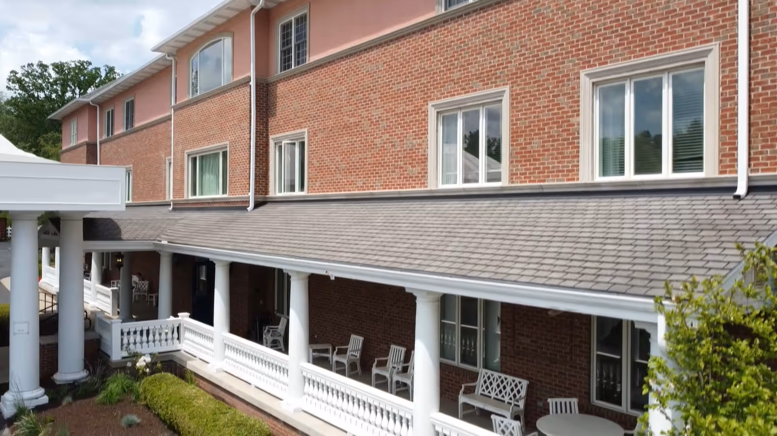 Exterior view of a brick senior living facility building with white columns supporting a covered porch area. Several white chairs and tables are arranged on the porch. Green shrubs and trees are visible around the building.