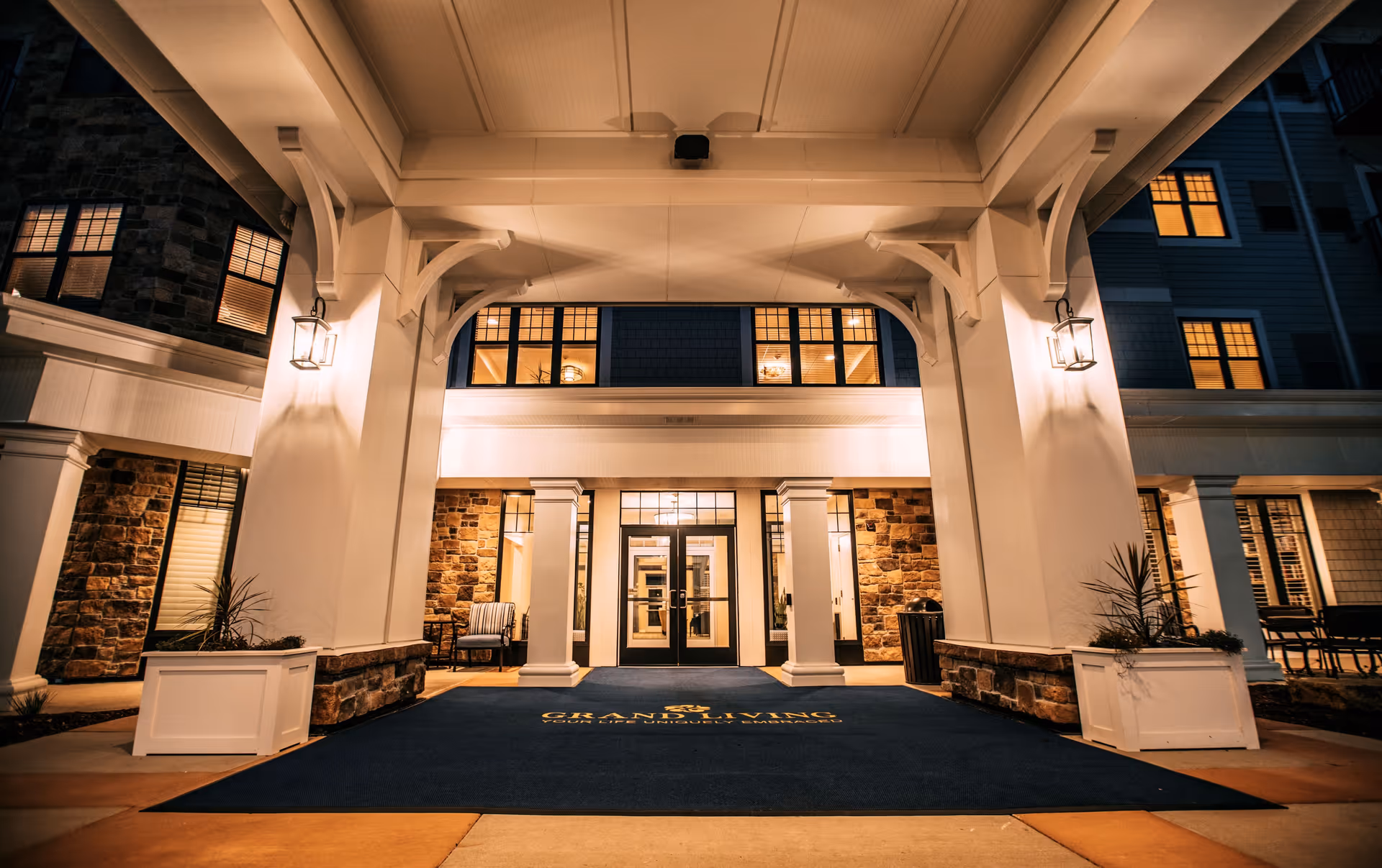 Night view of the entrance to a senior living facility with large white pillars, stone walls, and warm lighting. The entrance has glass double doors and a blue mat with the text 'Grand Living Your Life Uniquely Embraced'.
