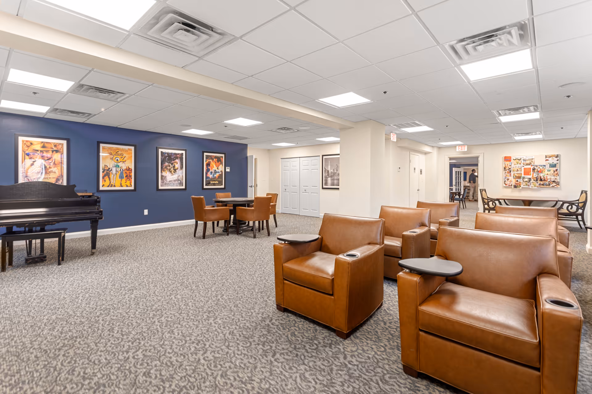 A spacious common area in a senior living facility with brown leather armchairs equipped with small attached tables and cup holders. In the background, there is a black grand piano against a blue accent wall adorned with framed movie posters. There are also a few tables with chairs and a bulletin board on the far wall. The room has a carpeted floor and a white ceiling with recessed lighting.