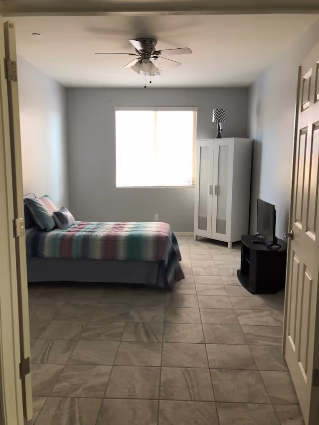 A minimal bedroom with a single bed covered in striped bedding, a white wardrobe, a small TV on a stand, a window, tiled floor, and a ceiling fan.