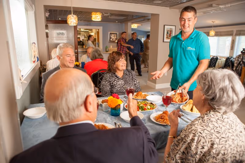 A group of elderly people sitting around a dining table enjoying a meal together in a well-lit room. A staff member in a teal shirt is serving spaghetti to the group. The table is set with plates of food, glasses of red juice, and a vase with yellow and orange flowers. Other people are visible in the background, engaging in conversation.