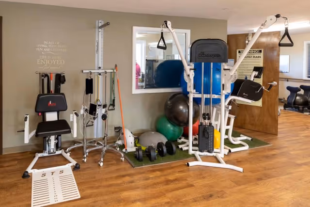 Small therapy/exercise room with a weight machine, exercise balls, free weights, and rehabilitation equipment on wood flooring.