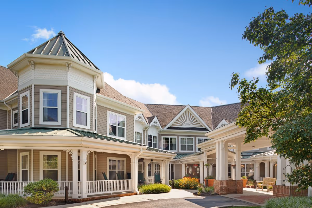 Exterior view of a senior living facility building with beige siding, white trim, multiple windows, and a covered entrance. The building features a turret-like structure and decorative architectural details. There are shrubs and trees around the entrance area under a clear blue sky.