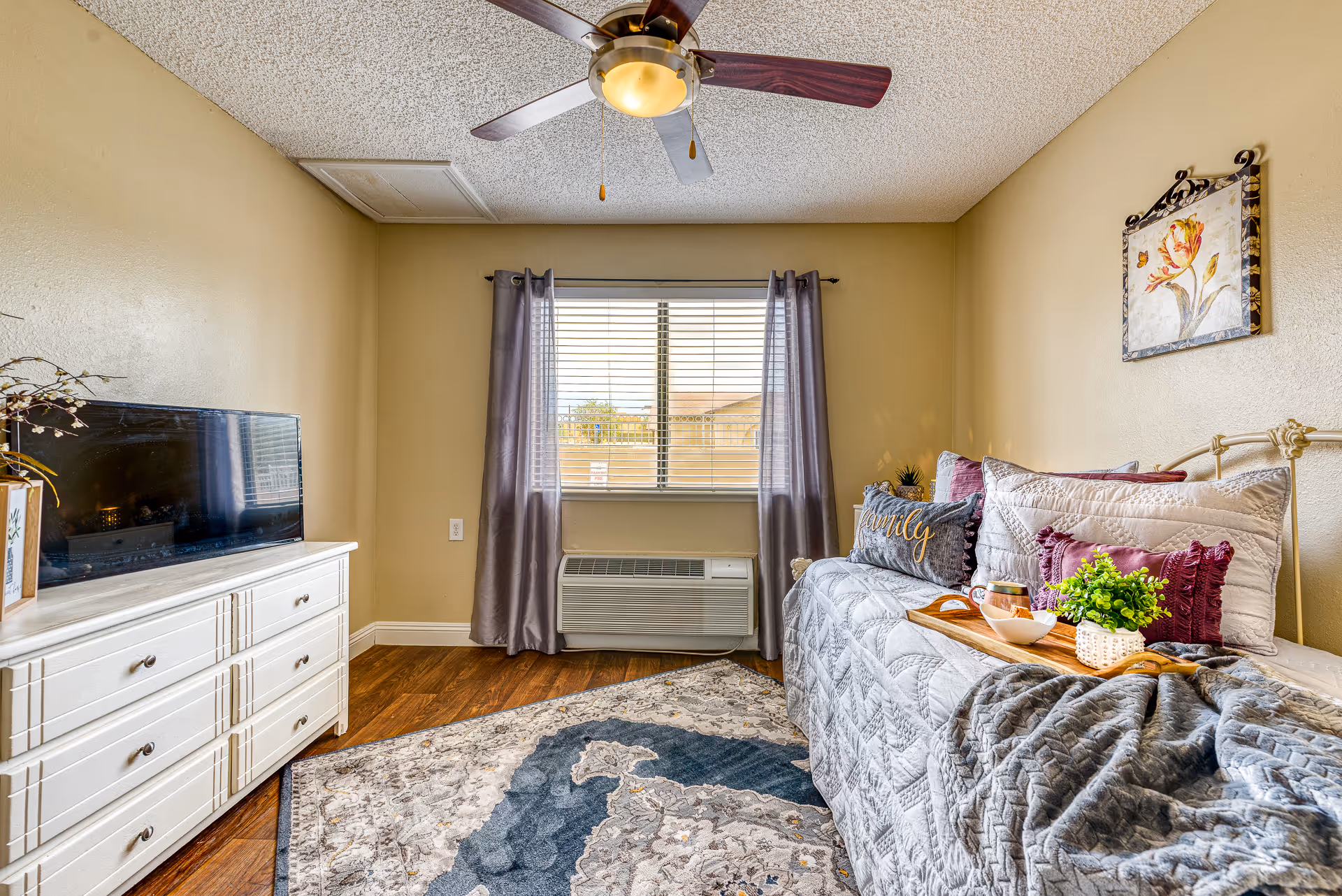 A cozy bedroom with a single bed covered in a gray quilt and multiple decorative pillows, including one with the word 'family'. A wooden tray with a cup, bowl, and small plant is placed on the bed. There is a window with gray curtains and blinds, an air conditioning unit below the window, a white dresser with a flat-screen TV on top, and a ceiling fan with wooden blades. The walls are painted beige, and a floral painting hangs above the bed. A patterned area rug covers part of the wooden floor.
