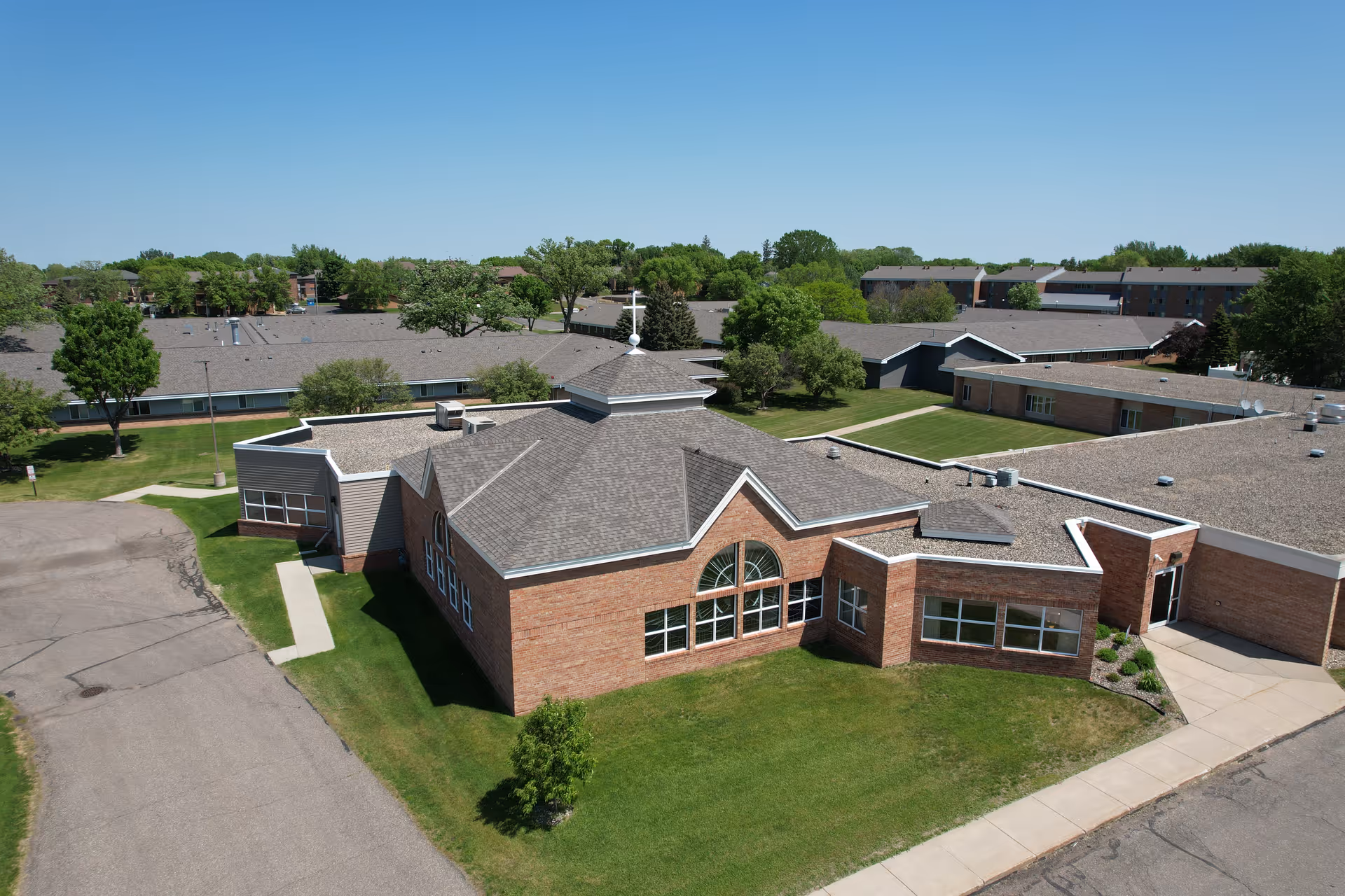 Aerial view of a senior living facility with multiple connected brick buildings, green lawns, trees, and paved walkways under a clear blue sky.