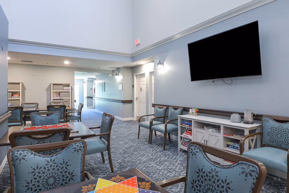 A bright assisted-living common area with multiple tables and patterned chairs, a wall-mounted TV, shelving with games, and a hallway in the background.