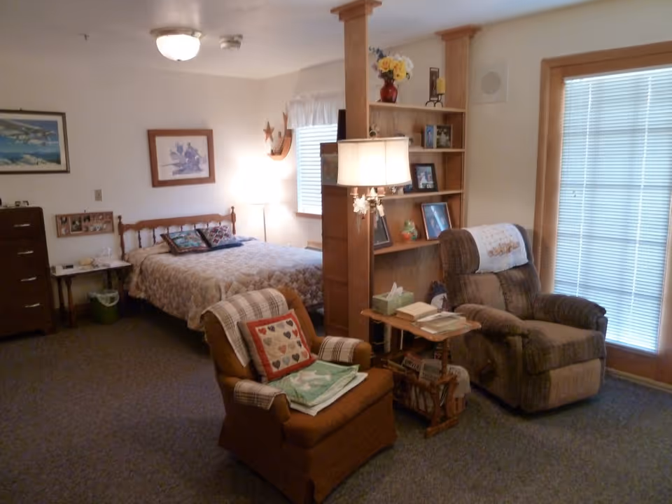 A cozy bedroom with a double bed covered in a quilt, two armchairs with cushions and blankets, a wooden bookshelf with framed photos and decorative items, a small side table with books and tissues, and a window with blinds letting in natural light.