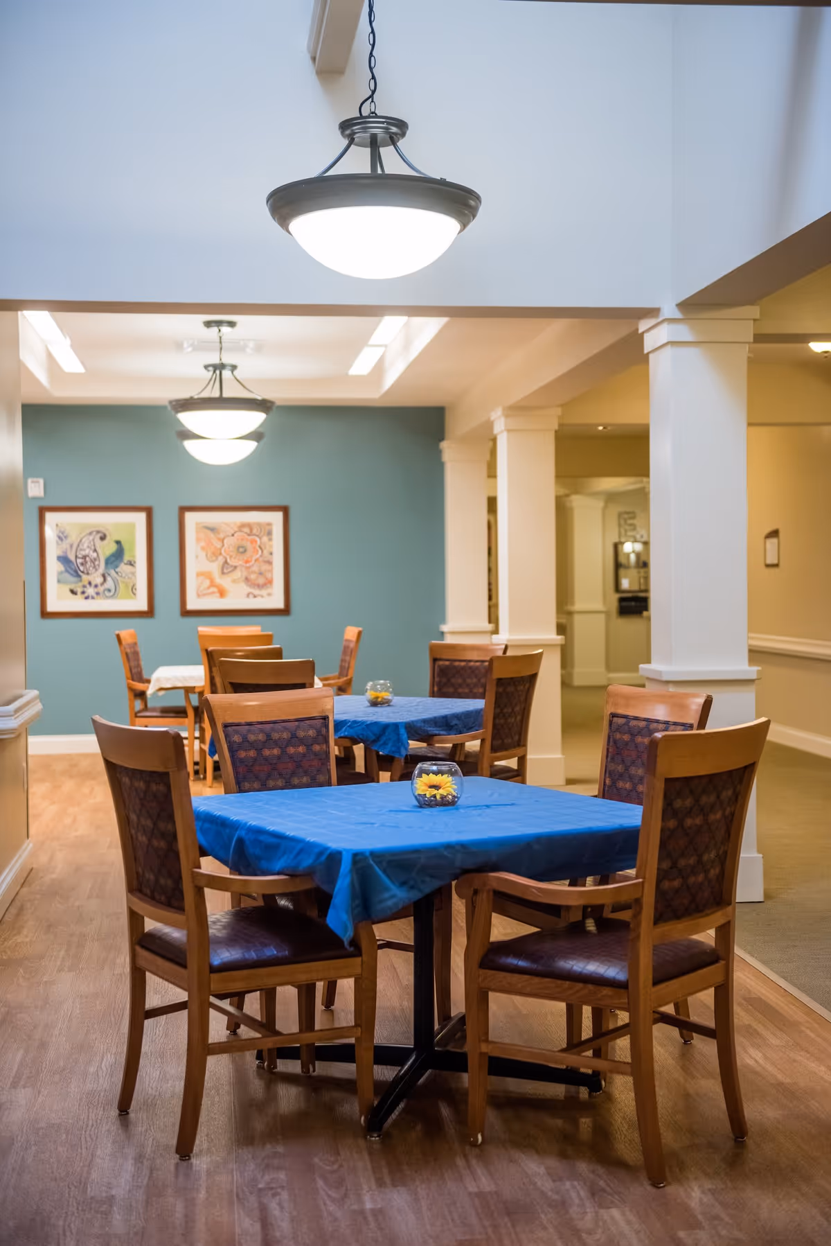 Interior view of a dining area in a senior living facility with wooden tables covered in blue tablecloths and wooden chairs. The room has light wood flooring, teal and beige walls, framed artwork, and hanging ceiling lights.