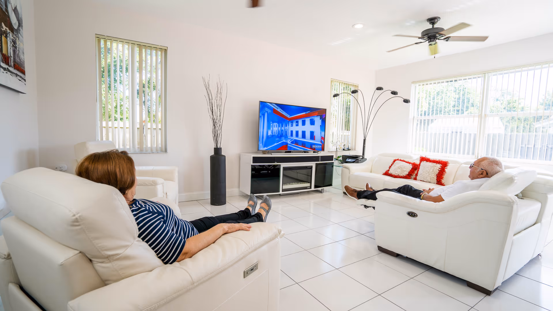 Two elderly people sitting on white leather recliners in a bright living room watching a flat-screen TV. The room has white tiled floors, large windows with vertical blinds, a ceiling fan, a modern floor lamp, and a TV stand with decorative items.
