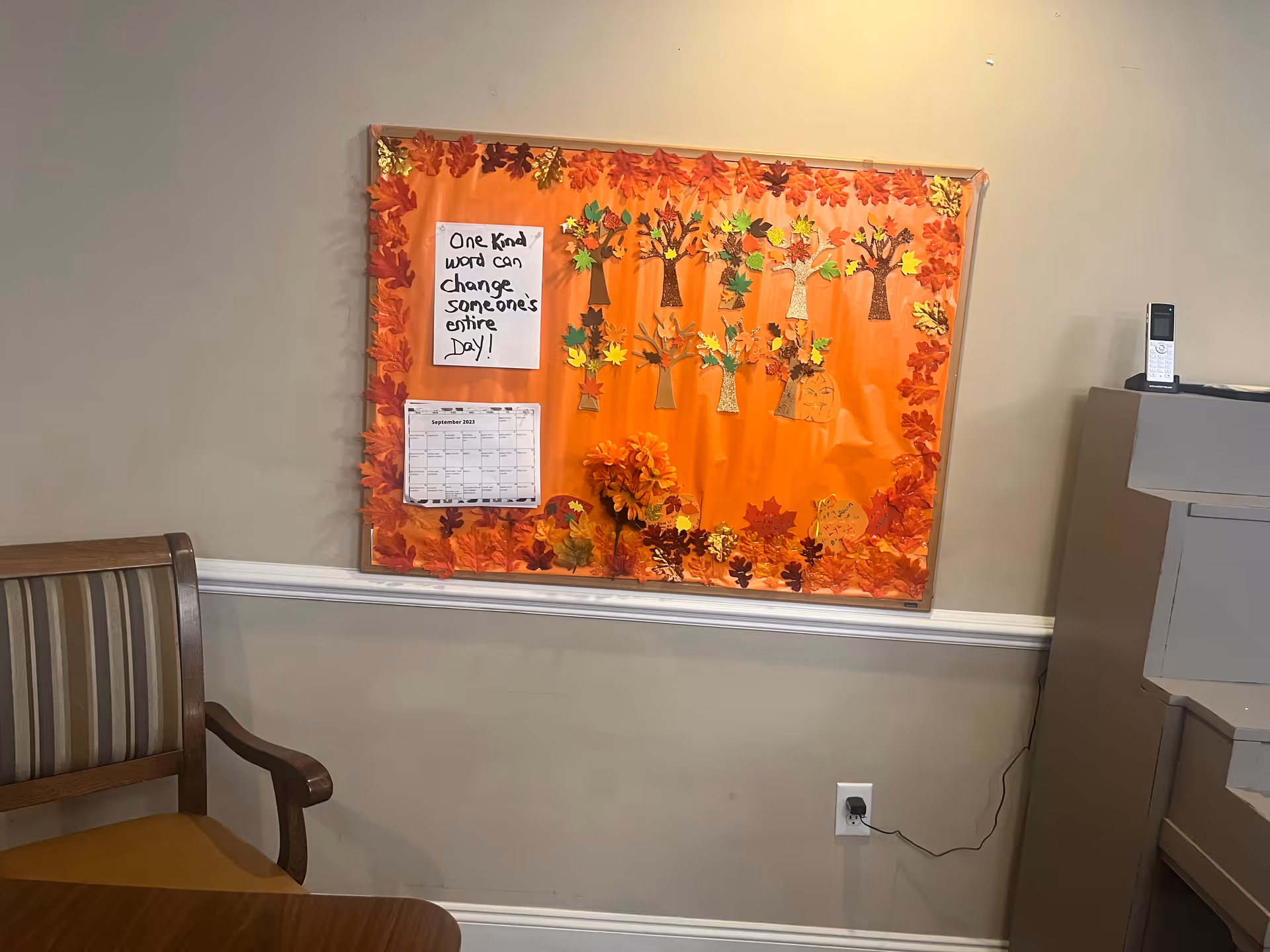 Indoor bulletin board decorated with orange autumn leaves and crafts mounted on a wall above a chair and table in a facility common area.