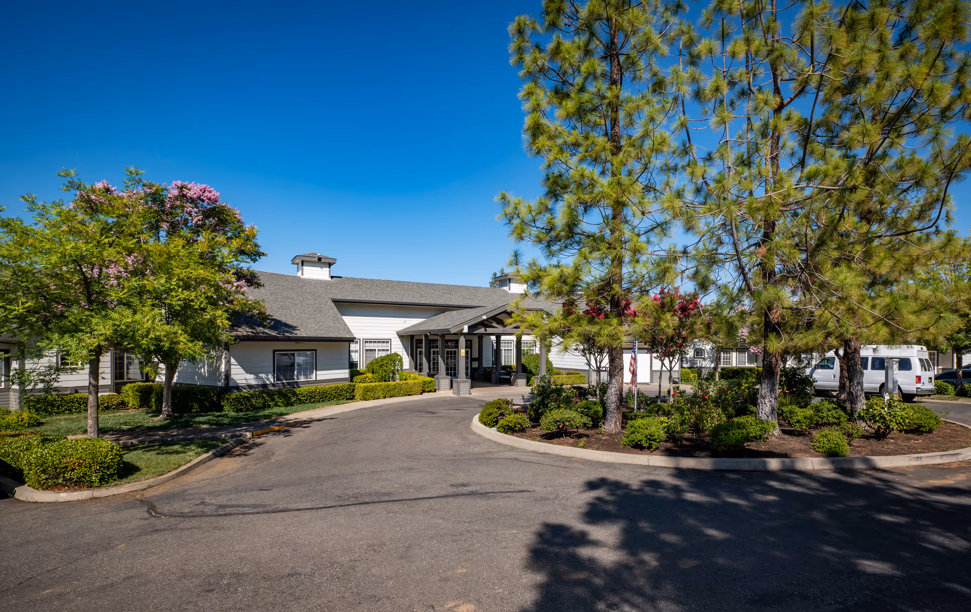 Exterior view of Country Crest Post-Acute facility showing a single-story building with a covered entrance, surrounded by landscaped greenery including trees and bushes under a clear blue sky.