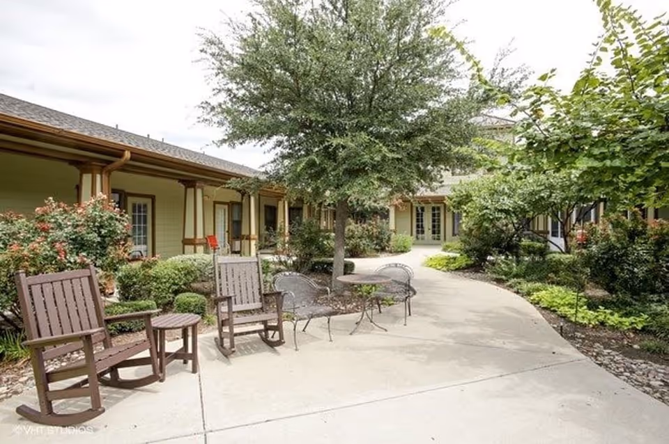 Outdoor courtyard area of Buffalo Creek Assisted Living and Memory Care featuring a paved walkway, wooden rocking chairs, metal chairs and tables, surrounded by green trees and shrubs with a building in the background.
