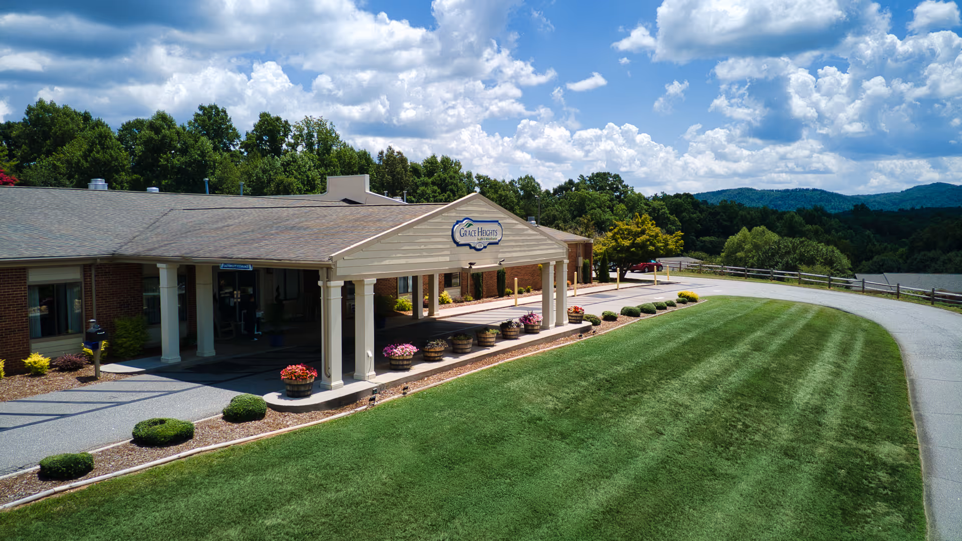 Front entrance of Grace Heights Health and Rehabilitation with a covered porte-cochère, flower planters, manicured lawn, and wooded hills in the background.