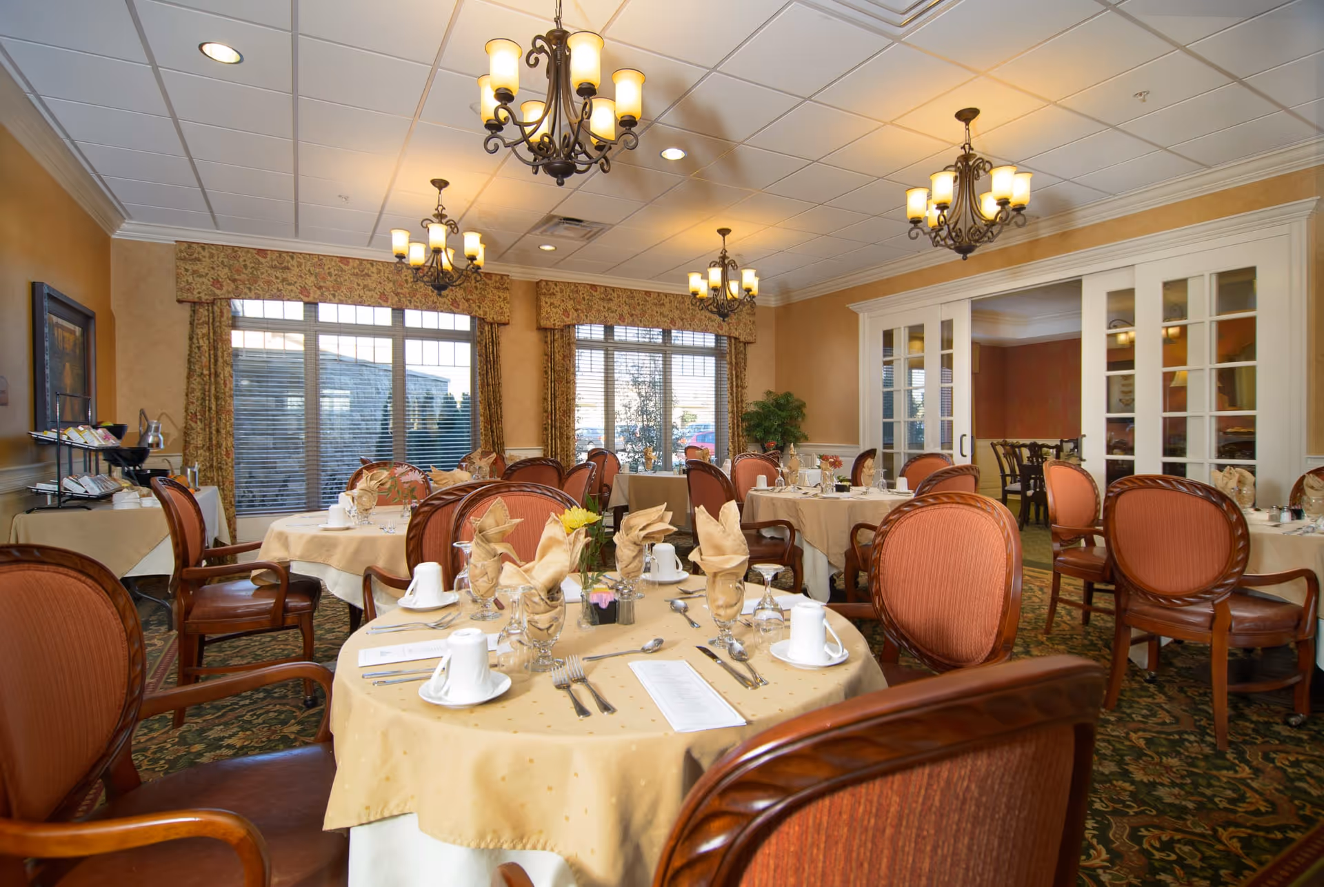 A well-lit dining room with round tables covered in beige tablecloths, each set with folded napkins, glassware, white cups, and silverware. The room features large windows with floral curtains, ornate chandeliers hanging from a white ceiling, and wooden chairs with cushioned seats. There is a side table with additional dining items and a plant in the corner.