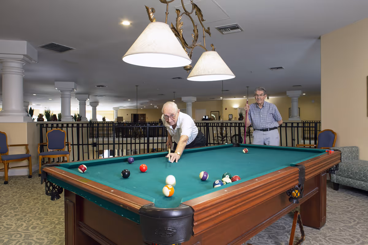 Two elderly men playing pool on a billiards table in a spacious senior living community game room.