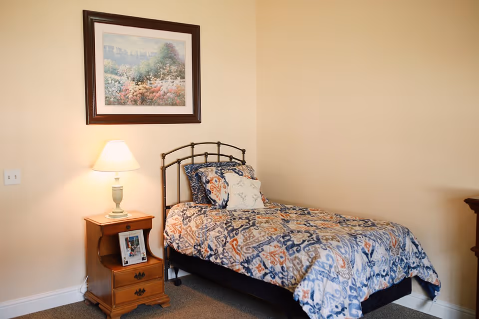 A small bedroom with a single bed covered in a patterned quilt, a wooden nightstand with a lamp and framed photo, and a framed floral painting on the wall.