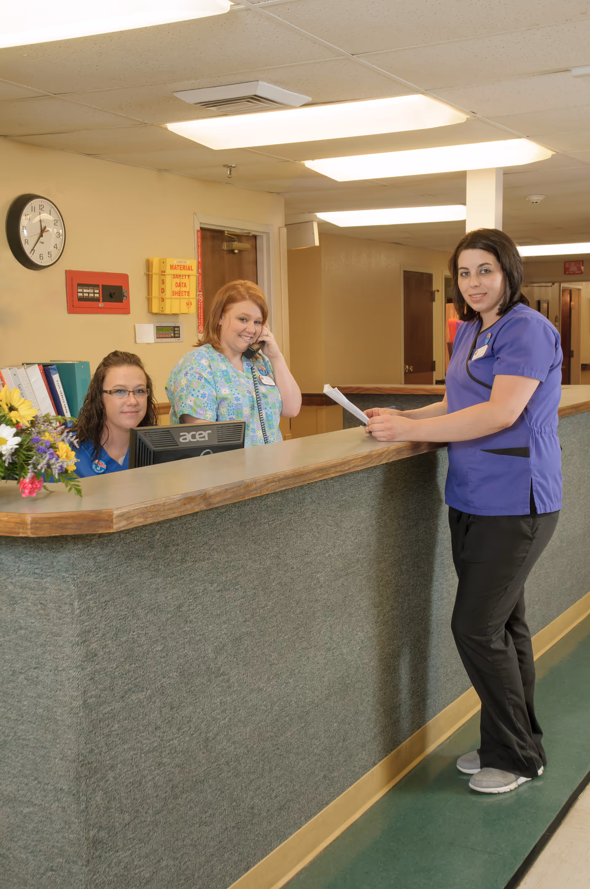 Three female healthcare workers at a reception desk in a senior living facility. One woman in purple scrubs is standing and holding papers, while two women behind the desk, one on the phone and one seated at a computer, are looking towards the camera. The area has beige walls, a clock, and office supplies visible.