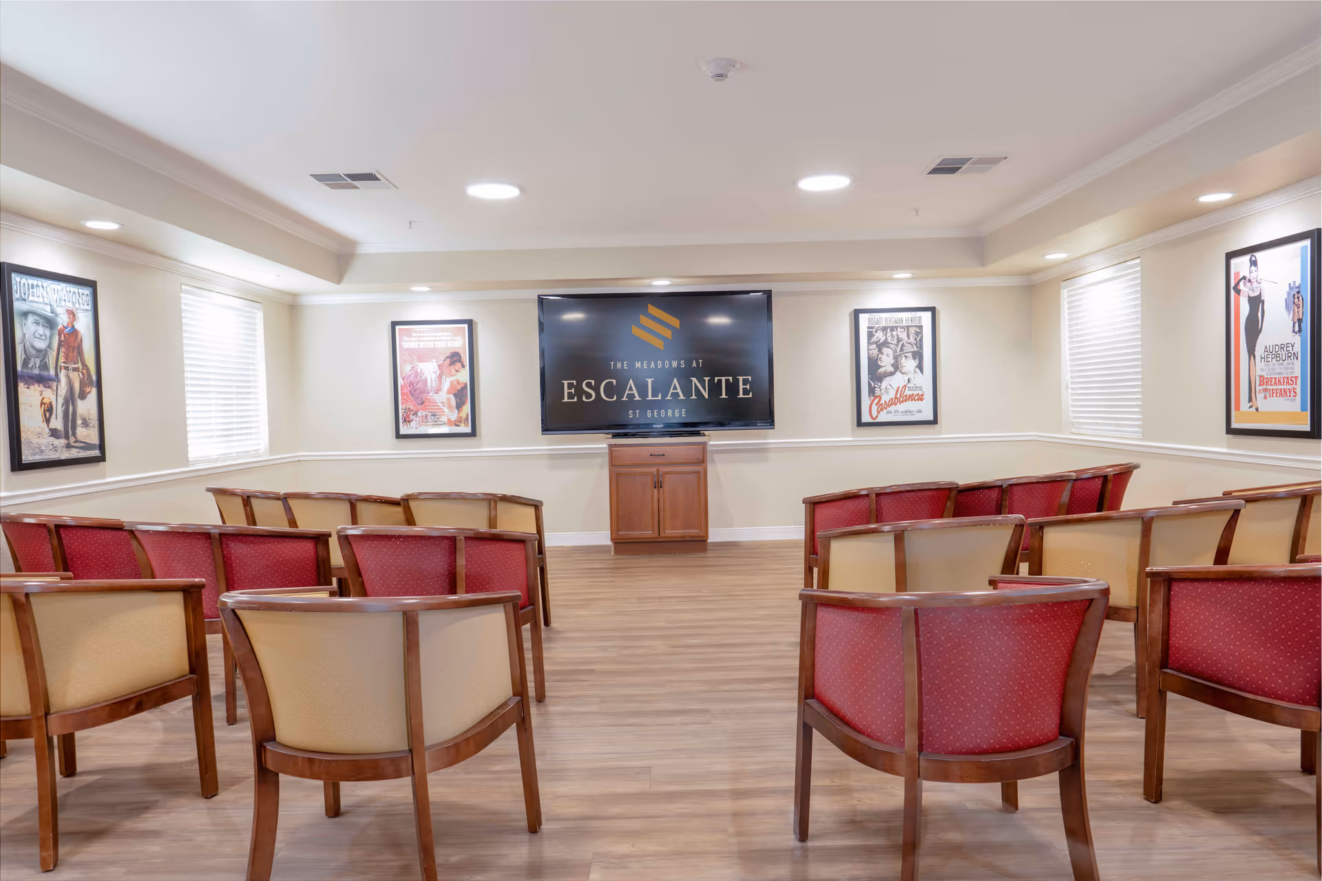 A small theater or presentation room with rows of wooden chairs upholstered in red and beige fabric facing a large flat-screen TV mounted on the wall. The TV displays the logo and name 'The Meadows at Escalante'. The room has light-colored walls, wooden flooring, and framed classic movie posters on the walls. Two windows with blinds allow natural light into the room.