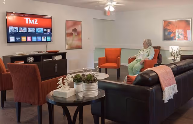 A senior woman sitting on an orange armchair in a living room area with a black leather sofa, orange chairs, a round table with plants, and a wall-mounted TV displaying the TMZ channel.