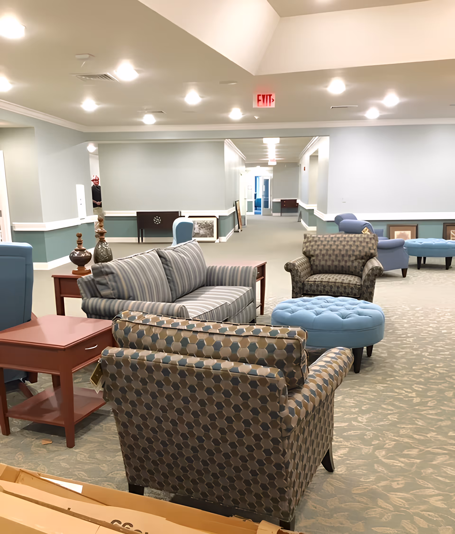 A spacious, well-lit common area in a senior living facility featuring multiple upholstered armchairs and sofas arranged around a blue tufted ottoman. The walls are painted light blue with white trim, and the carpet has a subtle floral pattern. The hallway extends into the background with framed pictures resting against the walls and an exit sign visible on the ceiling.