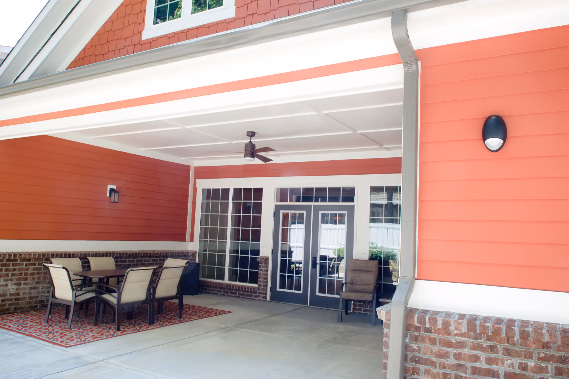 Covered outdoor patio area with a ceiling fan, a table with six chairs on a patterned rug, and a single chair near double glass doors. The walls are painted coral with white trim and brick accents.