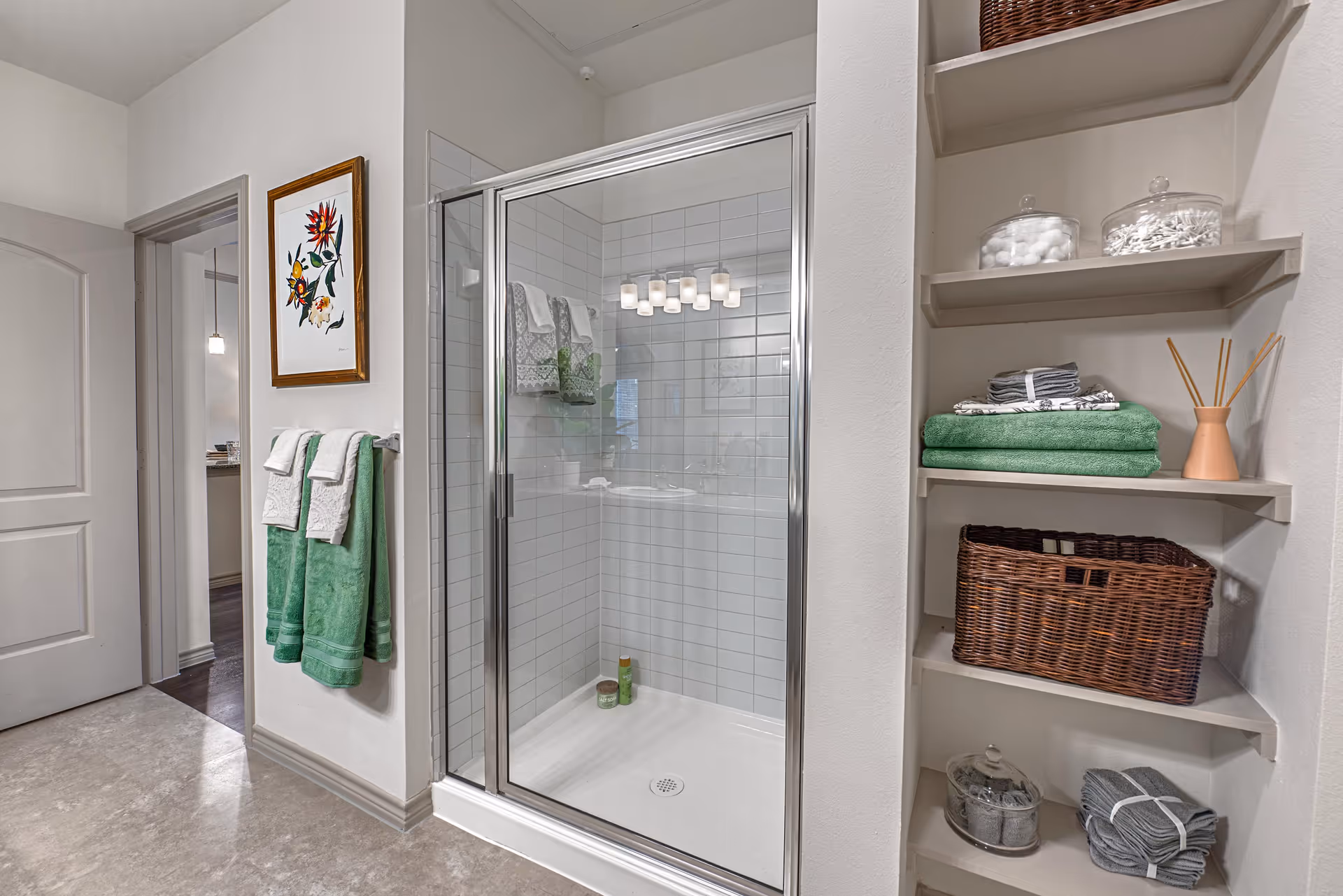 Glass-enclosed tiled shower in a bathroom with green towels on a rack and open shelves holding folded towels and wicker baskets.