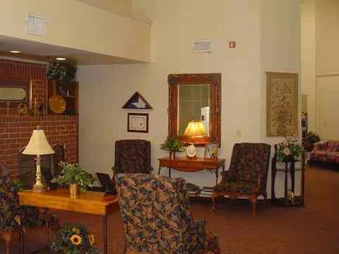 A cozy senior living lounge with patterned chairs, tables, lamps, and a brick fireplace.