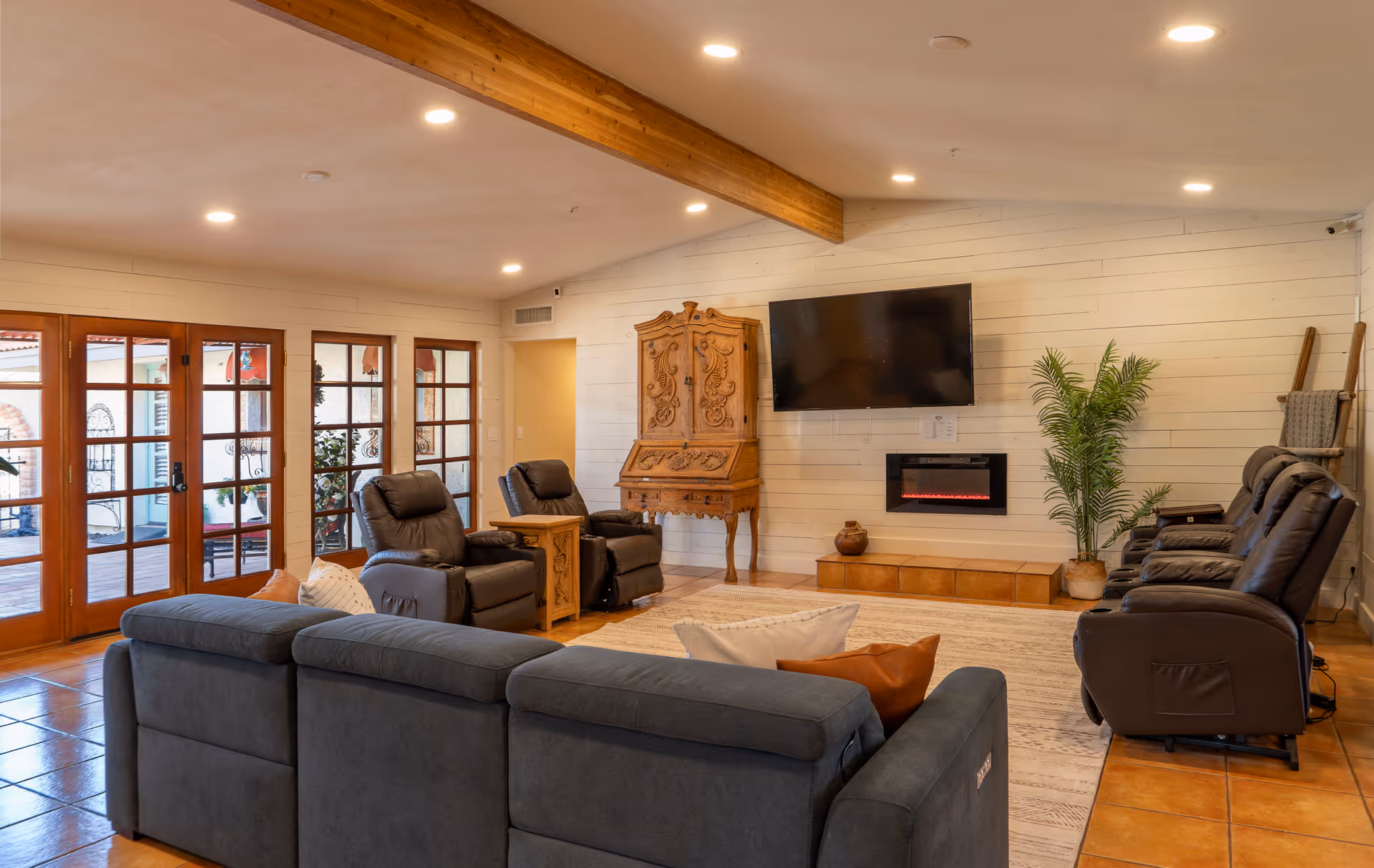A cozy living room with a large dark gray sectional sofa, two recliners with a wooden side table between them, and three additional recliners facing a wall-mounted flat screen TV above a modern electric fireplace. The room features wooden beams on the ceiling, white shiplap walls, a large area rug, and glass doors leading to an outdoor patio. There is also a decorative wooden cabinet and a potted plant in the corner.